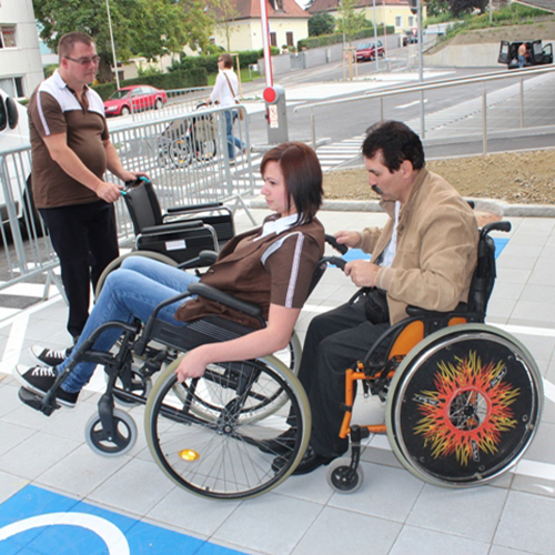 Zwei Personen im Rollstuhl, ein Mann ist behilflich